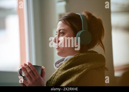 Femme réfléchie écoutant de la musique à travers des écouteurs sans fil enveloppés dans une couverture tenant tasse à café à la maison Banque D'Images