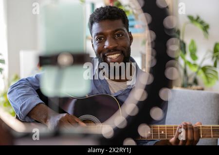 Heureux influenceur jouant de la guitare et filmant à travers un téléphone intelligent à la maison Banque D'Images