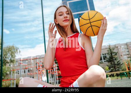 Joueur de basket-ball adolescent assis avec la balle sur le sol dans la cour d'école Banque D'Images