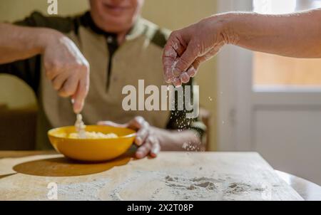 Homme senior mélangeant la pâte dans un bol et femme saupoudrant la farine sur la table Banque D'Images