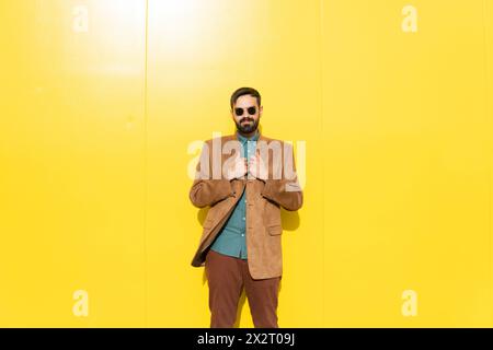 Homme souriant portant une veste marron et des lunettes de soleil devant le mur jaune Banque D'Images