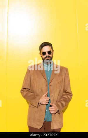Homme souriant portant une veste marron et des lunettes de soleil près du mur jaune Banque D'Images