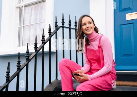 Heureuse belle femme portant une salopette rose avec téléphone intelligent assis sur les marches devant la porte bleue Banque D'Images