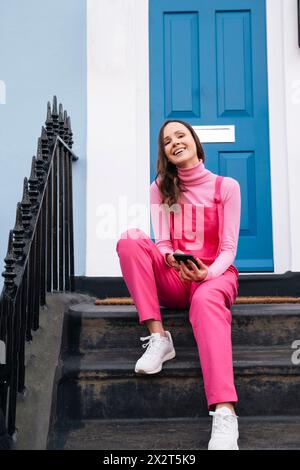 Femme heureuse portant salopette rose avec téléphone intelligent assis sur les marches devant la porte bleue Banque D'Images
