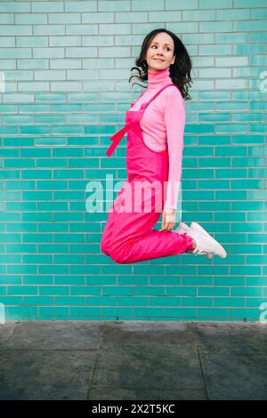 Femme souriante portant une salopette rose sautant devant un mur de briques bleues Banque D'Images