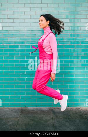 Femme portant une salopette rose sautant devant un mur de briques bleues Banque D'Images