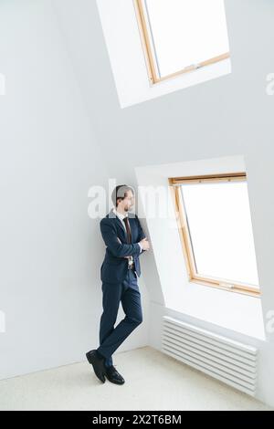 Homme d'affaires réfléchi avec les bras croisés penchés sur le mur blanc regardant par la fenêtre au bureau Banque D'Images