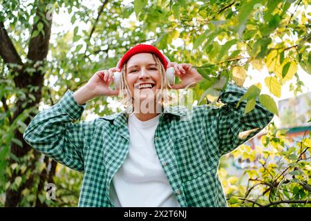 Jeune femme souriante écoutant de la musique avec des écouteurs sans fil et debout devant des arbres Banque D'Images