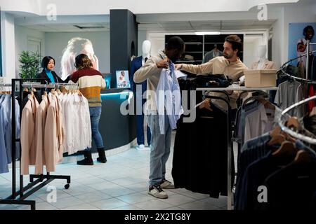 Client de magasin de vêtements essayant sur la chemise et obtenant des conseils de l'employé de boutique. Homme afro-américain tenant des vêtements au corps tout en choisissant la taille avec l'assistant de sortie de mode Banque D'Images