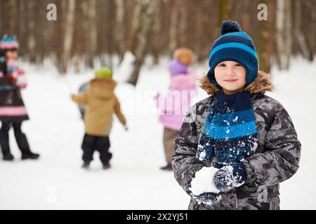 Portrait d'un garçon qui se tient dans le parc d'hiver et tient boule de neige dans ses mains, plusieurs enfants jouent derrière lui pas au point Banque D'Images