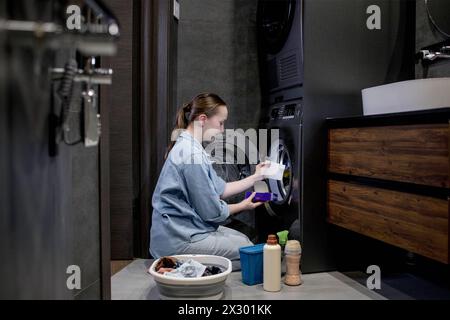 Femme assis devant une machine à laver chargeant des vêtements sales, serviettes pour laver des vêtements colorés. Banque D'Images