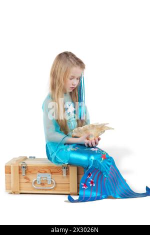 Petite fille habillée comme une sirène est assise sur la poitrine avec coquillage dans ses mains Banque D'Images