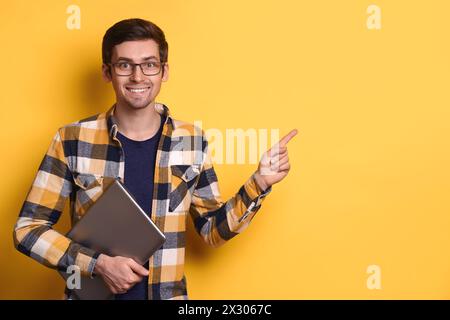 Intelligent jeune homme d'affaires, freelance, étudiant, programmeur, consultant tenant l'ordinateur portable, pointant l'espace de copie pour les mots clés, texte de publicité contre le mur jaune du studio. Concept de promotion et de publicité Banque D'Images