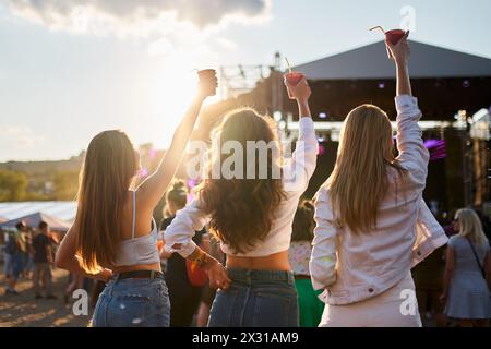 Groupe de femmes profiter de la musique à la fête de la plage, lever des boissons dans la célébration ensoleillée du bord de mer. Vue arrière, fête des femmes, danse près de la scène, ambiance amusante sur Banque D'Images