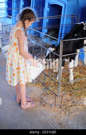 Heureuse petite fille tient un seau d'eau pour veau à la ferme de vache le jour d'été. Banque D'Images