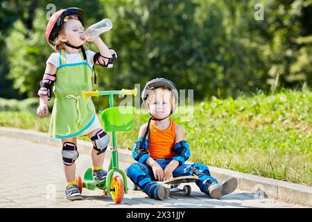 Le petit garçon est assis sur le skateboard à côté de la petite fille, qui boit de l'eau de la bouteille en plastique, debout avec un scooter à trois roues Banque D'Images