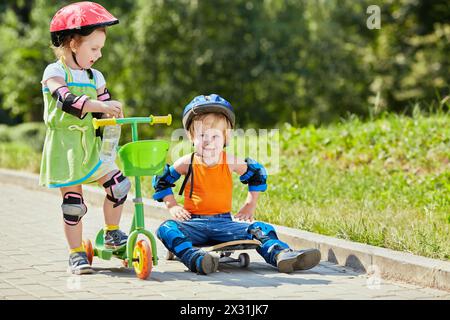 Petit garçon est assis sur le skateboard avec ses bras akimbo, petite fille avec trois roues scooter se tient à côté de lui Banque D'Images