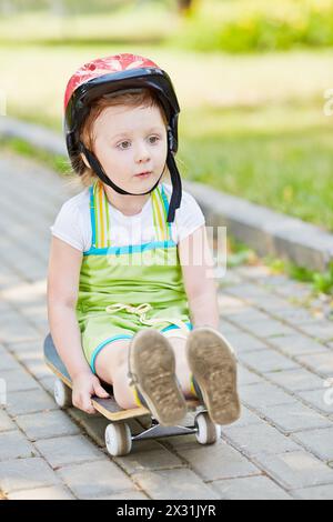 Petite fille dans un casque de protection descend l'allée du parc assise sur le skateboard Banque D'Images