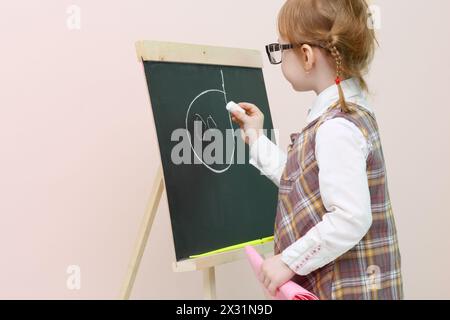 Petite fille avec des pigtails rouges dans des lunettes craie dessine le visage au tableau noir dans le studio. Banque D'Images