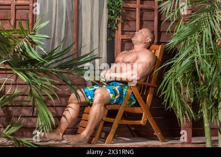 Homme musclé se reposant dans une chaise en bois à côté de la maison Banque D'Images