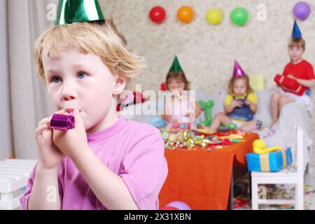 Petit garçon souffle dans des souffleurs de fête à la fête d'anniversaire et trois filles assis sur le canapé. Inscription joyeux anniversaire sur le mur. Concentrez-vous sur Boy. Banque D'Images