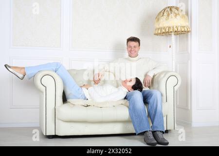 Le mari souriant est assis sur le canapé en cuir blanc et sa femme est couchée sur ses genoux à la maison. Banque D'Images