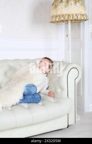 Petite fille dort sous la peau blanche sur un canapé blanc dans une pièce blanche à la maison. Banque D'Images