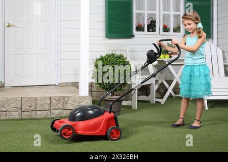 Joyeuse petite fille mignonne avec tondeuse à gazon rouge à côté de la maison blanche. Banque D'Images