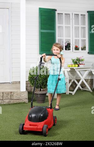 Joyeuse petite fille mignonne tond la pelouse par tondeuse à gazon rouge à côté de la maison blanche. Banque D'Images