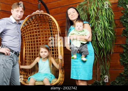 Petite fille est assise dans une chaise suspendue en osier et père, mère avec support de bébé près d'elle. Banque D'Images