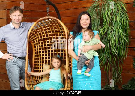 Petite fille est assise dans la chaise suspendue en osier et père, mère avec support de bébé près du mur en bois. Banque D'Images