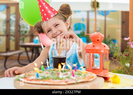 Petite fille dans un chapeau de fête dans un café avec une pizza et une boisson Banque D'Images