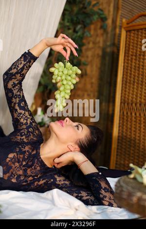 Belle femme se trouve sur le lit et tient un bouquet de raisins verts dans la chambre. Banque D'Images