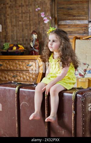 La petite fille aux pieds d'ours en robe jaune est assise sur une vieille grosse valise déchiquetée Banque D'Images