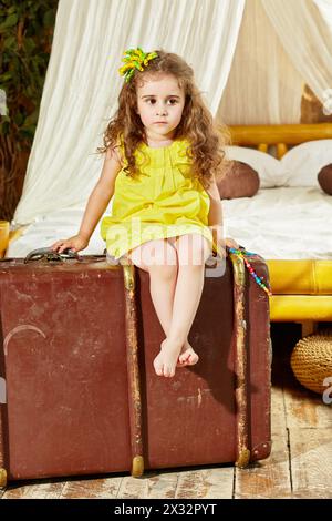 La petite fille en robe jaune est assise sur la vieille grande valise déchiquetée dans la chambre avec un grand lit sous la tente Banque D'Images