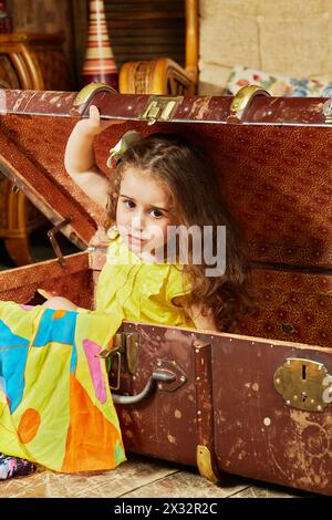 La petite fille en robe jaune sort de la vieille grosse valise en fibre déchiquetée couchée sur le sol dans la chambre Banque D'Images