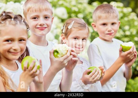 Les enfants se tiennent contre des buissons en fleurs tenant des pommes vertes à moitié mangées, concentrez-vous sur le garçon deuxième à gauche Banque D'Images