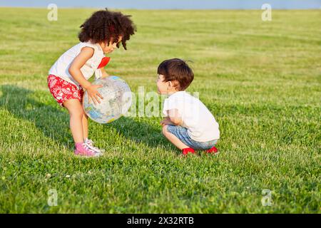 Petit garçon et petite fille jouent avec ballon-globe gonflable sur prairie herbeuse, la fille tient la balle, le garçon est assis squatté Banque D'Images