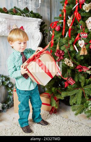 Le petit garçon se tient près de l'arbre de Noël, tenant une boîte en carton attachée avec un ruban rouge Banque D'Images