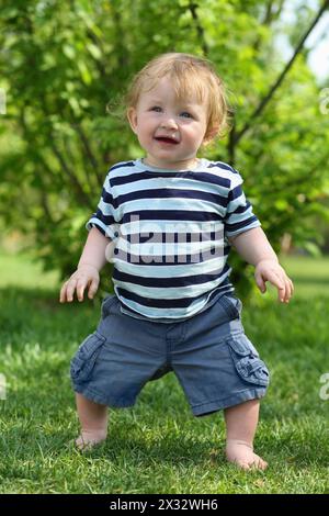 Petit garçon pieds nus en T-shirt rayé se tient sur l'herbe et à la journée ensoleillée en plein air. Banque D'Images