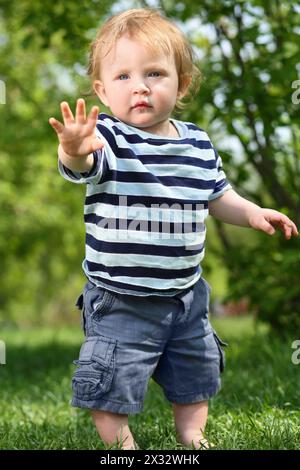 Petit garçon pieds nus en short se tient sur l'herbe verte et tend la main à la journée ensoleillée. Banque D'Images