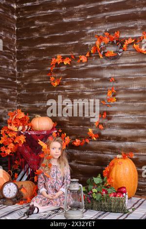 Petite fille est assise sur le sol avec une vieille lampe et un panier de fruits dans la chambre avec des citrouilles et un intérieur d'automne. Banque D'Images