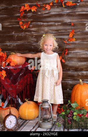 Petite fille se tient parmi la vieille lampe, panier de fruits, grandes citrouilles dans la chambre avec intérieur d'automne. Banque D'Images