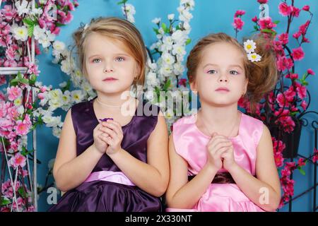 Deux petites filles mignonnes en robes s'assoient avec les mains pliées dans la prière dans la chambre avec des fleurs. Banque D'Images
