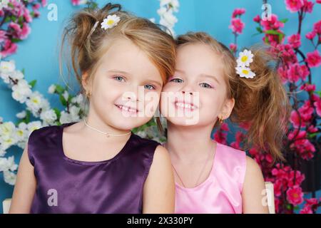 Deux petites filles mignonnes en robes regardent la caméra et sourient dans la chambre avec des fleurs. Banque D'Images