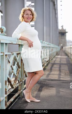 Heureuse belle femme enceinte en robe blanche se tient sur le pont. Banque D'Images