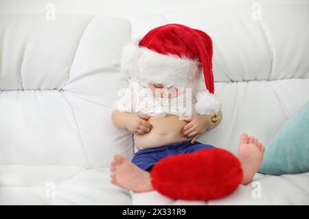 Mignon petit enfant dans le chapeau rouge est assis sur le canapé blanc et regarde son ventre à la maison. Faible profondeur de champ. Banque D'Images