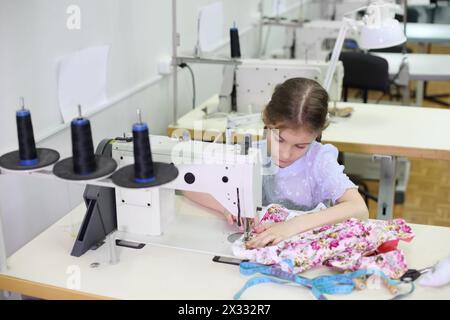 La fille étudiante coud une petite robe à la machine à coudre blanche dans la salle de classe avec de nombreuses machines. Banque D'Images