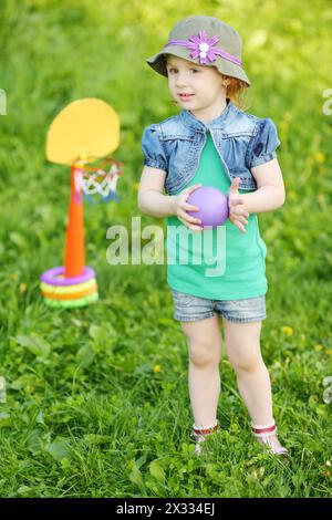 Petite jolie fille tient le ballon et se tient près du mini panier de basket-ball à Green Meadow. Banque D'Images