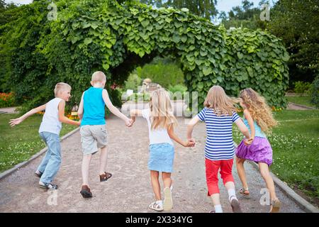 Les enfants courent en tenant les mains sur la passerelle du parc dans l'arc fait de plantes vertes, vue arrière, se concentrer sur les filles au centre Banque D'Images